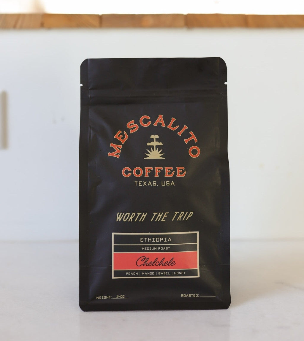 A black bag of Mescalito Coffee’s Single Origin Whole Coffee Beans | Chelchele sits on a white surface; this medium roast features Ethiopian beans (anaerobic natural process) with tasting notes of peach, mango, basil, and honey.