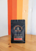 A black bag of Mescalito Coffee’s Single Origin Whole Coffee Beans | Madriz stands upright on a wooden table, with an orange and white wall in the background. The medium roast beans are sourced from Madriz, Nicaragua.