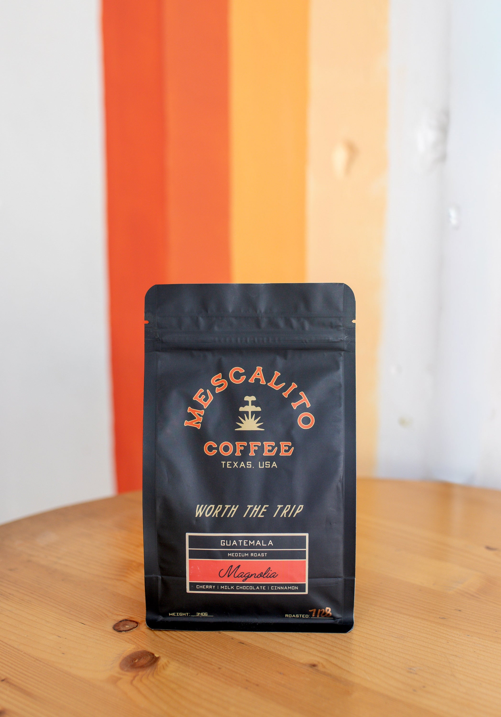 A black bag of Mescalito Coffee’s Magnolia Single Origin Whole Coffee Beans sits on a wooden table, set against a white and orange striped wall, showcasing its expertly crafted medium roast Guatemalan flavor.